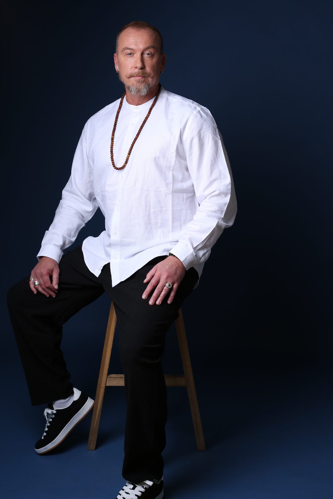 Ross Leslie Nicol seated on a stool in a white shirt with wooden mala beads